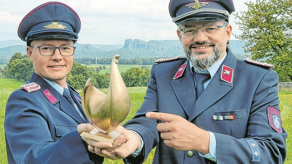 Die Feuerwehrmänner Toni Schulze (li.) und Kai Bigge mit der Goldenen Henne vor einem Panorama in der Sächsischen Schweiz.
